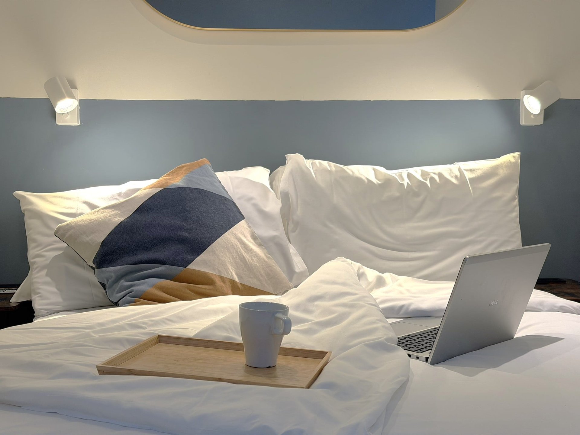 Laptop and wooden tray with mug on a bed.